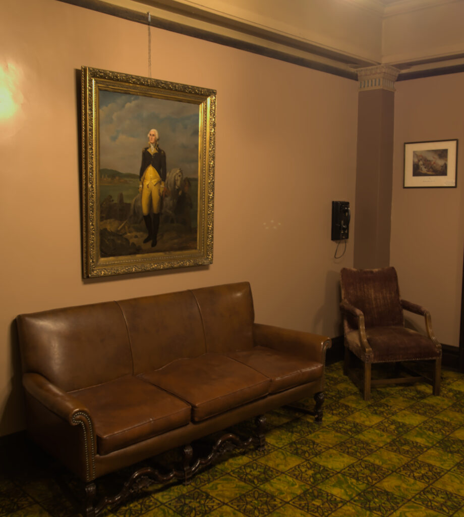 George Washington painting on the wall above a tired leather couch. There's also another old black phone on the wall next to a smaller worn velvet chair.