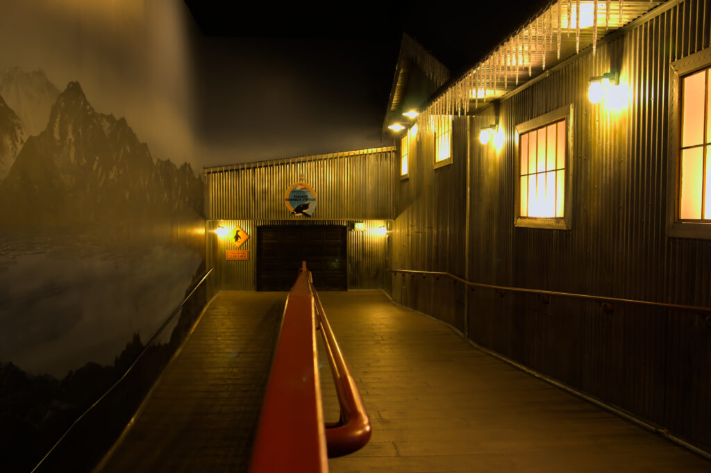 A hallway with decending slope. Red handrail with a flat top down the middle, old looking corrugated steel walls, fake windows, some old looking lights, and garage door at the turning point.